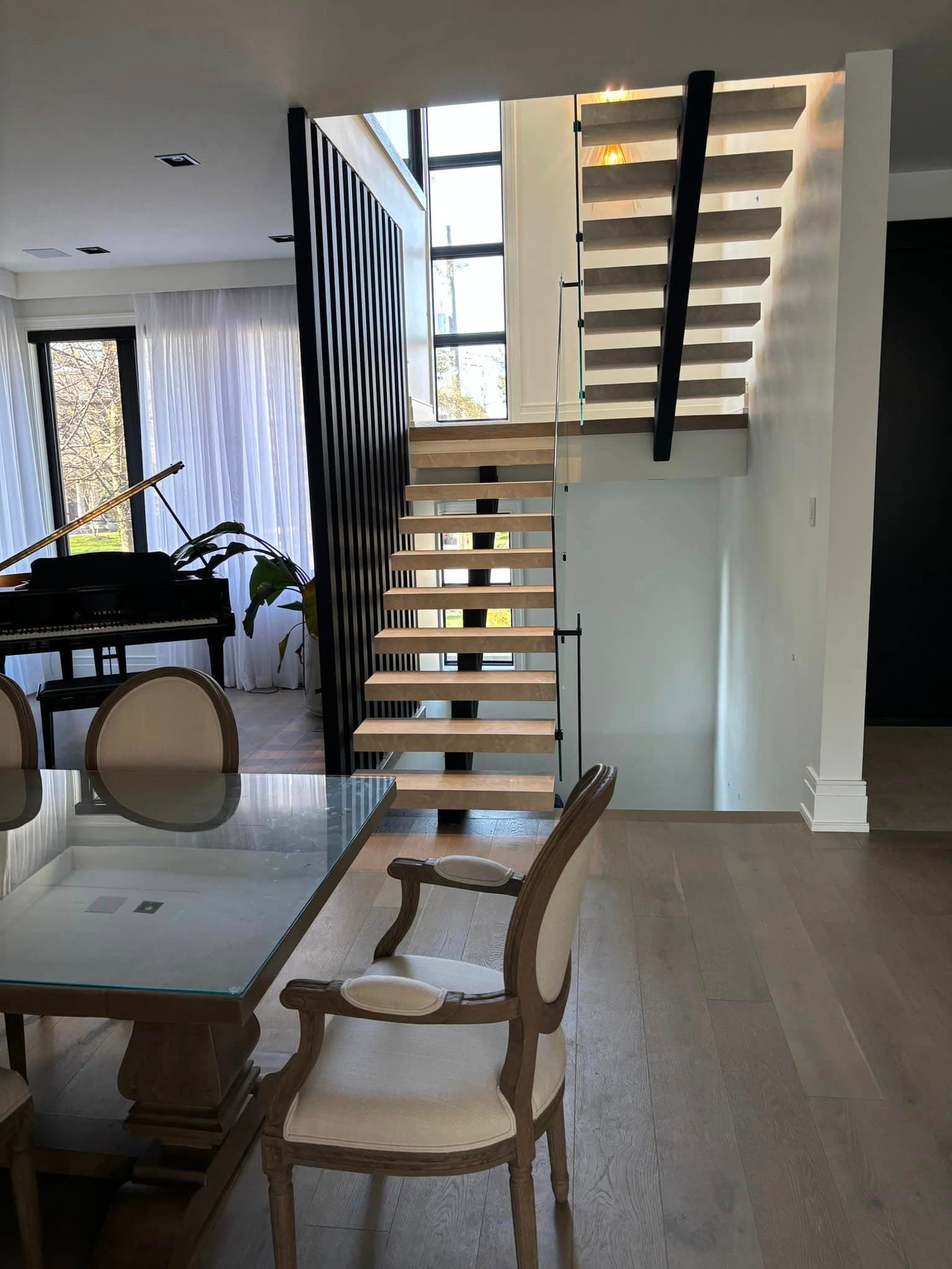 Modern open-plan room with a floating wooden staircase, grand piano, and glass-top dining table.