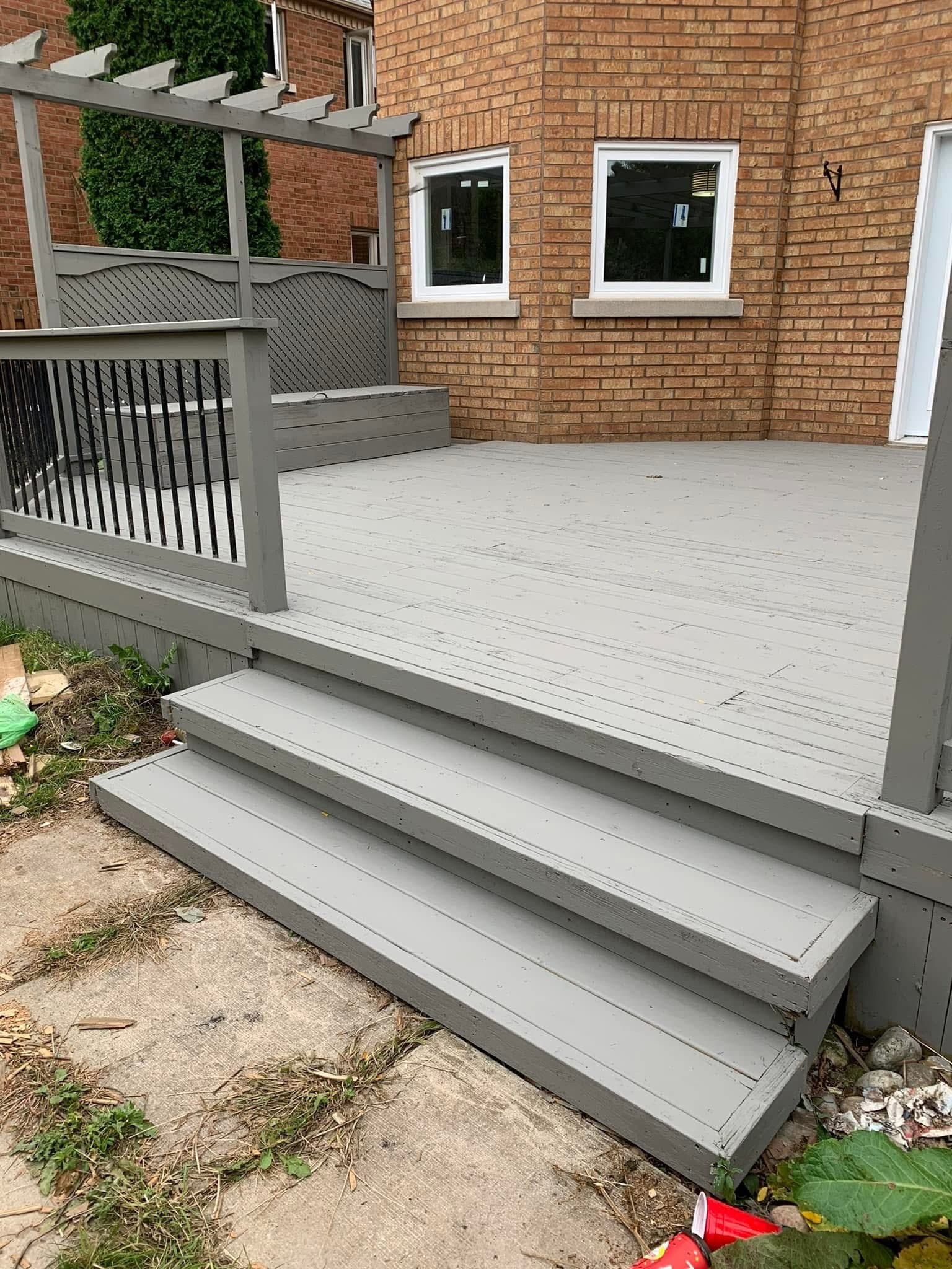 Grey painted wooden deck with wide steps, railing, and pergola against a brick house.