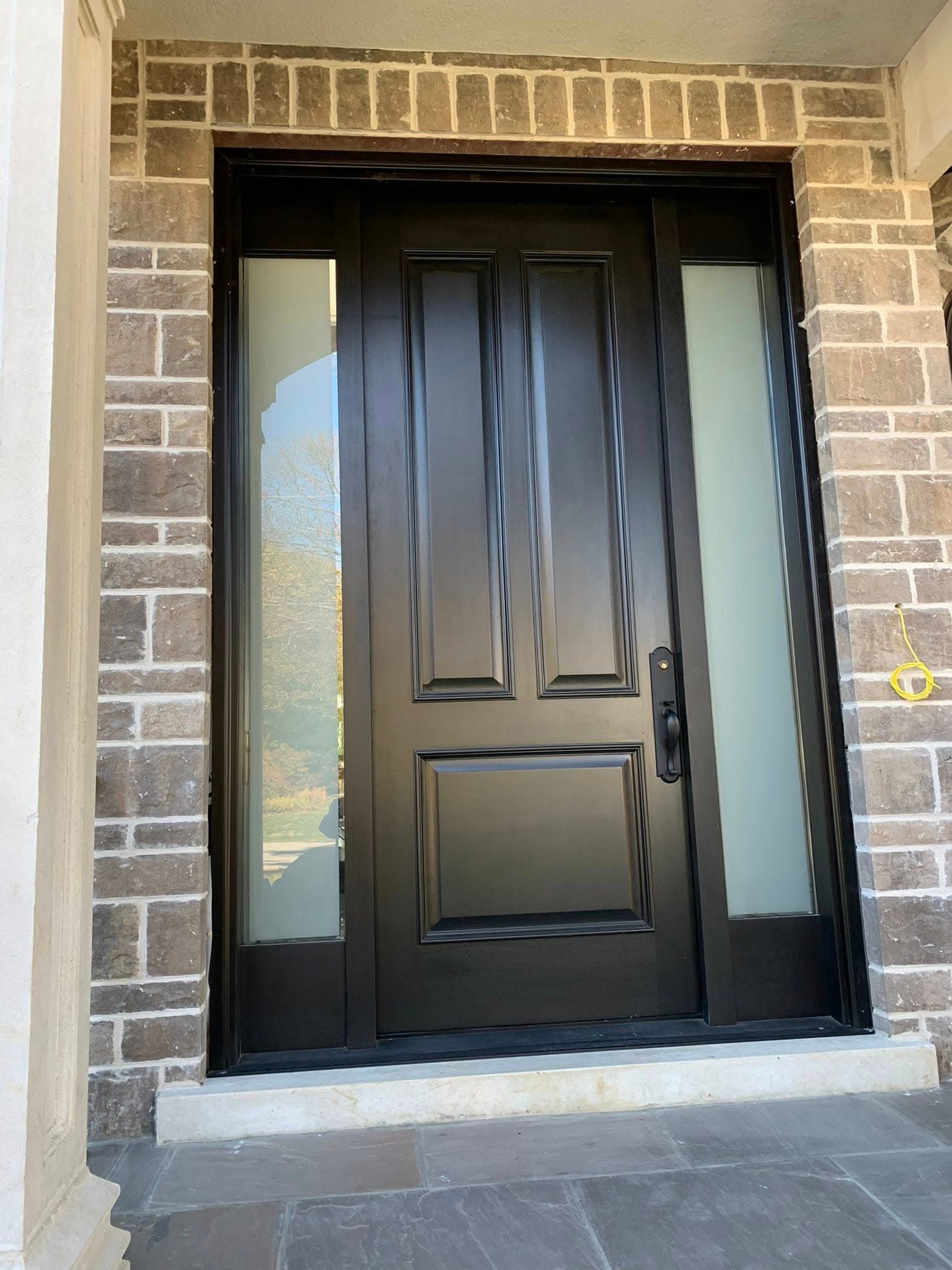 Black three-panel front door featuring frosted glass sidelights and a light brown brick surround.