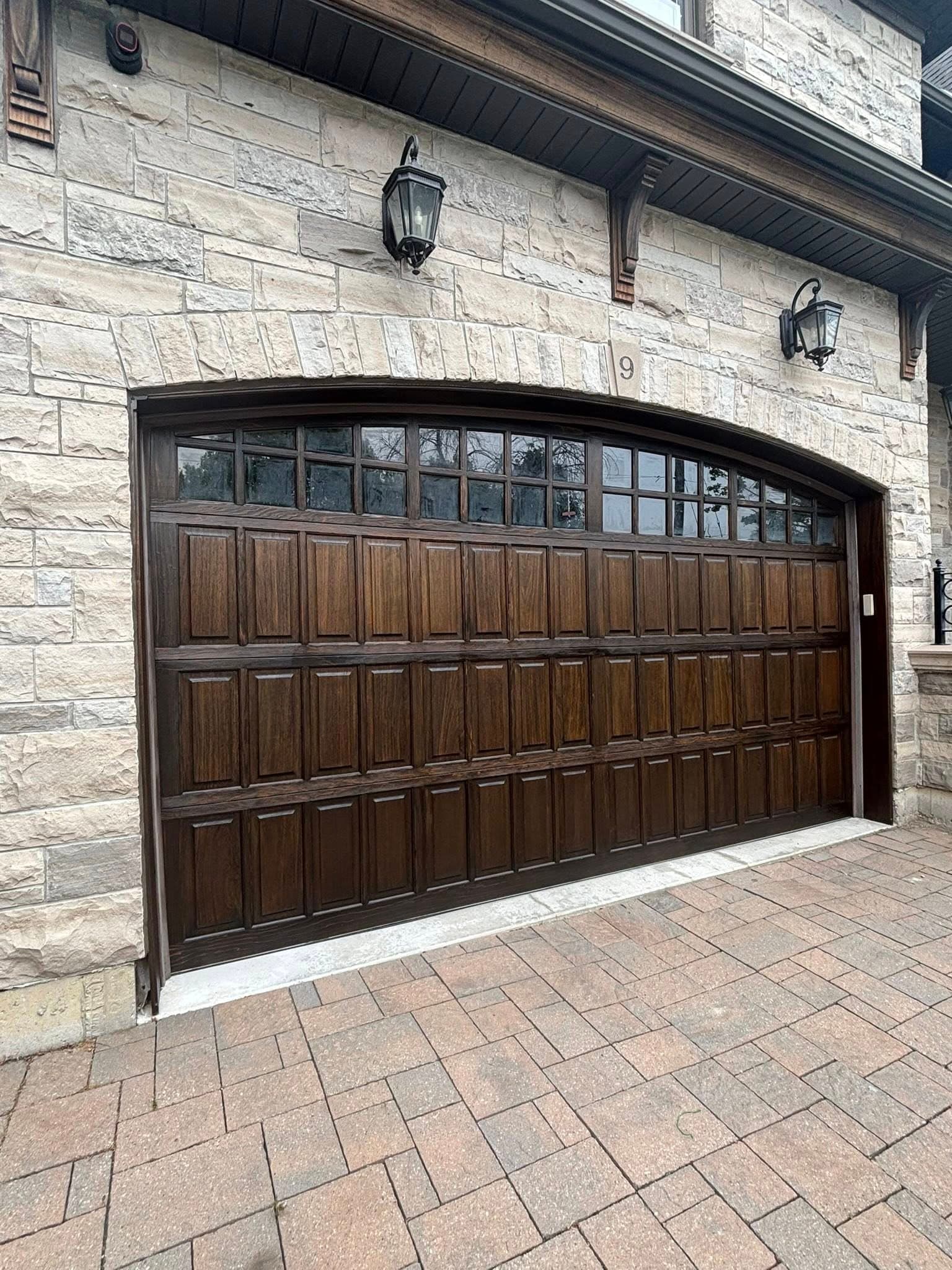 Arched dark wood garage door with windows, set in a light stone wall with lanterns.